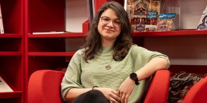 Die junge Frau sitzt vor einem roten Bücherregal in einem roten Sessel. Sie trägt einen hellgrünen Pullover, eine Brille und ihre braunen Haare offen. Den linken Arm hat sie auf der Armlehne. Sie lächelt mit geschlossenem Mund in die Kamera.