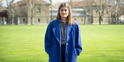 Eine Frau mit einem blauen Mantel und braunen Haaren steht vor einer grünen Wiese und schaut in die Kamera. Im Hintergrund sieht man Bäume ohne Blätter und Gebäude.