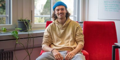 Der junge Mann trägt sein braunes Haar offen bis zum Kinn, ein blaues Fisherman-Beanie und einen gelbbeigen Hoodie. Er sitzt auf einem roten Kinosessel vor einem Fenster. Auf der Fensterbank steht eine Pflanze.