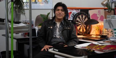 Der junge Mann trägt die Haare und  schwarze Jacke offen. Er sitzt an seinem Atelierplatz, eine Pultlampe brennt gelblich, eine Dartscheibe und viele Flyer und Werke sind zu sehen.