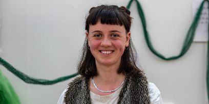 Porträtaufnahme der jungen Frau mit dunklem Haar. Sie trägt sie bis zu den Schultern, hat Fransen und einen Teil der Haare zu zwei Pompons am Hinterkopf hochgebunden. Sie trägt ein weisses Oberteil, darüber eine Art Fellweste und eine silberne Kette. Sie strahlt in die Kamera.