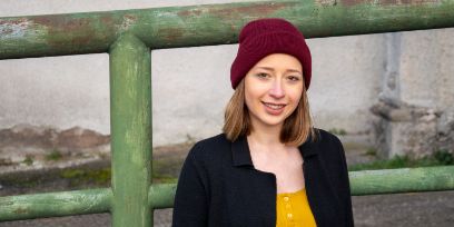 Die dunkelblonde Frau trägt eine beerenrote Mütze auf dem Kopf. Die Haare sind schulterlang offen zu sehen. Sie trägt ein senfgelbes Shirt mit feinen Knöpfen vorne und darüber ein schwarzes Jäckchen. Sie steht vor einem grossen grünen Rohrgeländer, das bereits etwas Rost oder darunterliegende rötlichbraune Farbe zum Vorschein kommt.