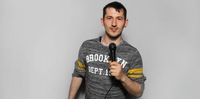 Der Mann steht mit Mikrophon in der linken Hand und offenem Blick in die Kamera schauend in der Mitte des Bildes mit leichter Neigung zu seiner Linken. Er trägt einen grau-melierten Pullover mit der Aufschrift BROOKLYN.