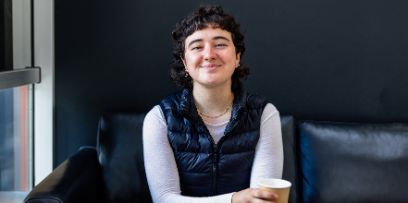 Portrait von Camille Santacroce. Die junge Frau sitzt auf einem schwarzen Sofa und blickt mit Kaffeebecher in der Hand direkt in die Kamera. Camille lacht.