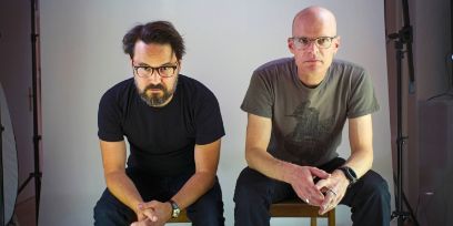 Die beiden Musiker sitzen auf Stühlen, beugen die Oberkörper leicht nach vorne sodass ihre Ellebogen auf ihren Oberschenkeln abstützen und sich die Hände jeweils berühren.