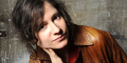 Die Frau vor einer Betonmauer mit geneigter Körperhaltung, in brauner Lederjacke mit halboffenem braunem Haar, die Finger ihrer rechten Hand an der rechten Wange, in die Kamera blickend