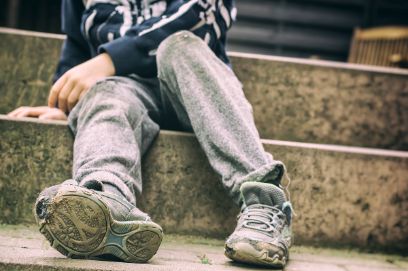 Eine Person sitzt auf einer Steintreppe. Die Person trägt Turnschuhe und eine Jogginghose. Der Turnschuh sieht abgenutzt aus und hat ein Loch bei den Zehen.