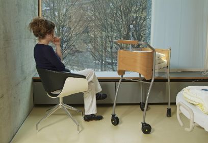 A woman is sitting by the window of a care home and looking outside. Next to her is a baby cot.