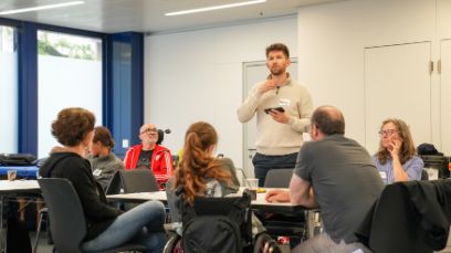 A young man is standing and explaining something to a group of people sitting down in a seminar room.