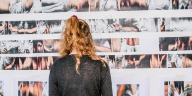 Eine Frau steht vor einer weissen Wand, die mit diversen Fotografien behängt ist. Die Frau hat helles, zusammengebundenes Haar und trägt einen schwarzen Pullover.