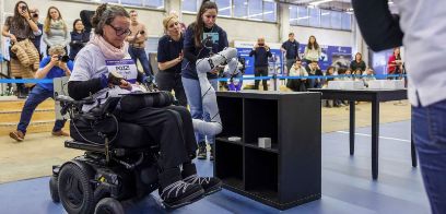 Frau in Elektrorollstuhl steuert Roboterarm und greift damit nach einem Gegenstand.