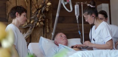 Foto: Pflegefachfrau untersucht einen Patienten mit Stethoskop, zwei weitere Pflegefachfrauen schauen zu.