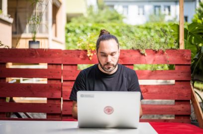 Max Frischknecht im Portrait, lange Haare zusammengebunden, Schwarzes Shirt, vor Laptop sitzend