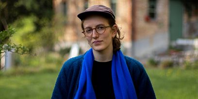 Die Musikerin steht vor einem Haus in grüner Umgebung. Sie trägt ein dunkelblaues Oberteil, einen knallblauen Schal, eine Brille und eine Schirmmütze