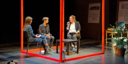 Drei Personen sitzen in einem roten Holzkonstrukt auf einer Bühne. Die Person rechts im Bild hält ein Mikrofon und spricht.