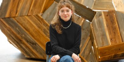 Die junge Frau sitzt vor einem Kunstwerk aus Holz. Sie trägt die dunkelblonden Haare halboffen, einen schwarzen Rollkragenpullover und Bluejeans. Die Beine hat sie überschlagen und die Arme ruhen überkreuzt auf den Beinen. Sie blickt lächelnd in die Kamera.