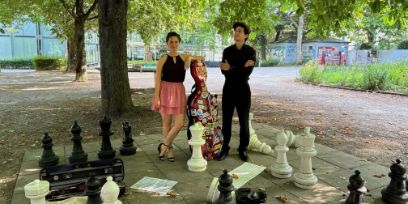 Das Duo «TangoSauvage» steht auf einem öffentlichen Schachbrett in einem Park. Es ist Sommer. Viviane Ruof lehnt mit ihrem linken Arm locker m Cello-Koffer ihres Partners, der links davon in schwarz mit verschränkten Armen steht..