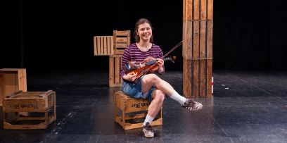 Eine junge Frau sitzt auf einer Bühne mit diversen Kisten aus Holz. Sie hat die Beine verschränkt und hält eine Violine in der Hand.