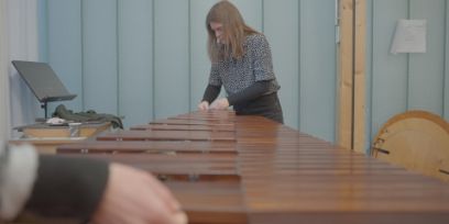 Eine Frau steht am Ende einer Marimba und arbeitet daran. Im linken Vordergrund ist unscharf eine Hand zu sehen.