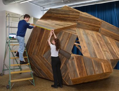 Links steht ein junger Mann auf einer Leiter, rechts davon eine junge Frau am Boden. Beide arbeiten an einem grossen Holzkonstrukt mit dem Namen «Halber Findling (Martin)».