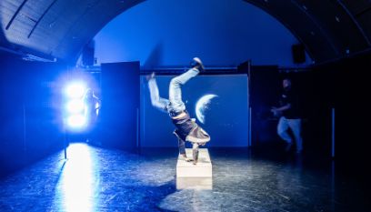 Ein einem blau ausgeleuchteten Raum mit Tonnendach macht ein Schauspieler in der Mitte einen Handstand. Im Hintergrund links schaute eine Frau zu, im Hintergrund rechts ein Mann.