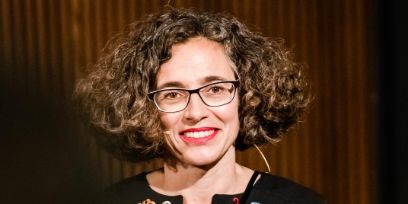 Porträt von Nina Mekacher: Sie lächelt und blickt in die Kamera, trägt roten Lippenstift und eine dunkle Brille. Ihr gelocktes, sehr voluminöses Haar trägt sie auf Kinnhöhe offen.