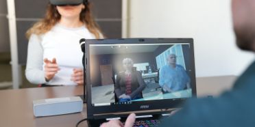 Eine Frau trägt eine VR-Brille. Sie sitzt einem Dozenten gegenüber.